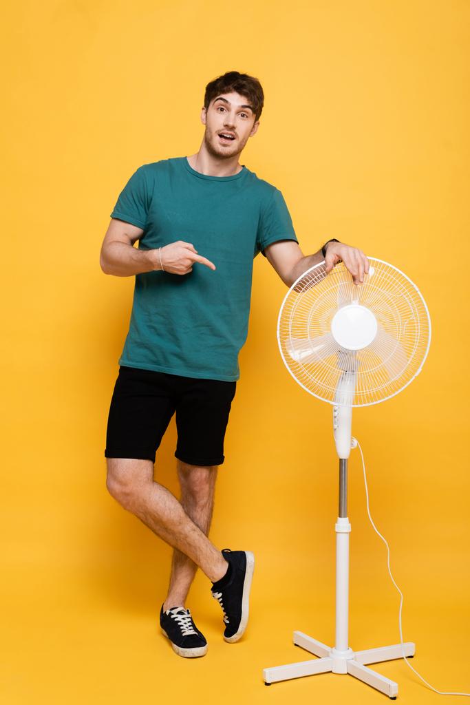 удивленный молодой человек показывает пальцем на желтый электрический вентилятор
     - Фото, изображение