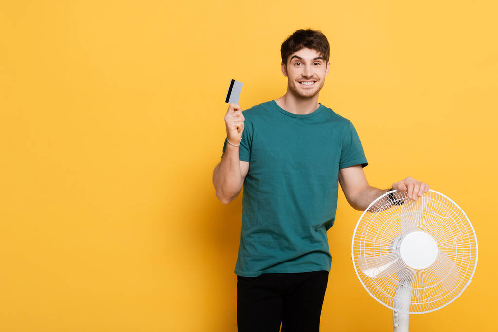 улыбающийся мужчина держит кредитную карту, стоя с электрическим вентилятором на желтом
      - Фото, изображение