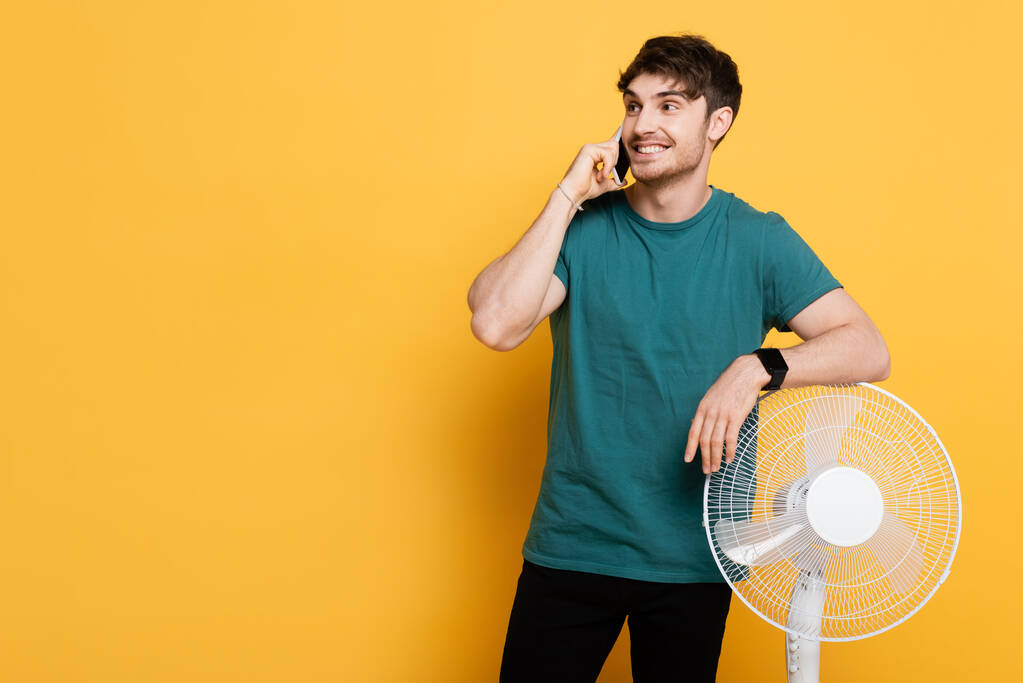 smiling man talking on cellphone while standing near electric fan on yellow      - Photo, Image