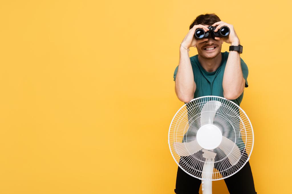 веселый человек смотрит через бинокль, стоя с электрическим вентилятором на желтом
      - Фото, изображение