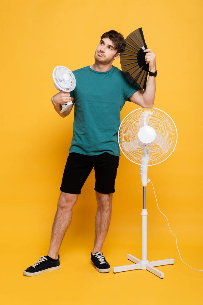 όμορφος άντρας που υποφέρει από ζέστη με δύο ηλεκτρικούς ανεμιστήρες και ανεμιστήρα χειρός στο κίτρινο    - Φωτογραφία, εικόνα