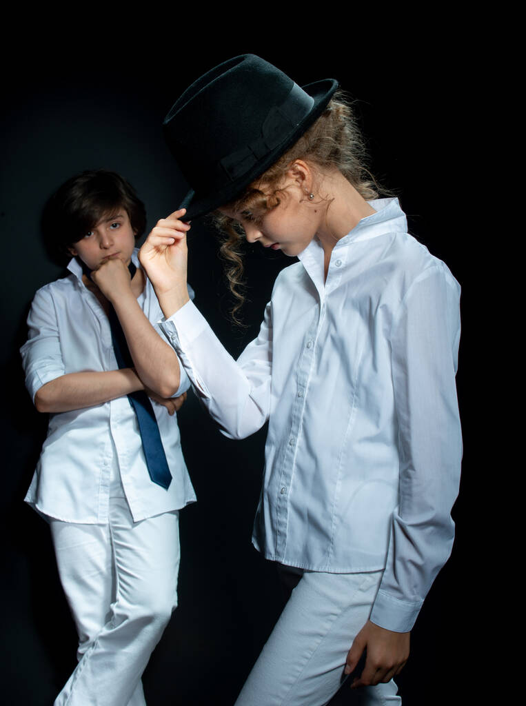 мальчик и девочка в белых рубашках и шляпе позируют в студии на темном фоне
 - Фото, изображение