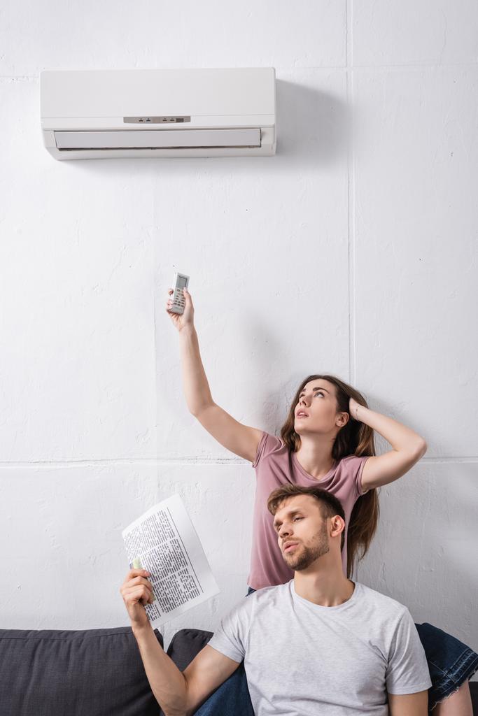 jovem casal com controlador remoto e jornal sofrendo de calor enquanto sentado em casa com ar condicionado quebrado
  - Foto, Imagem