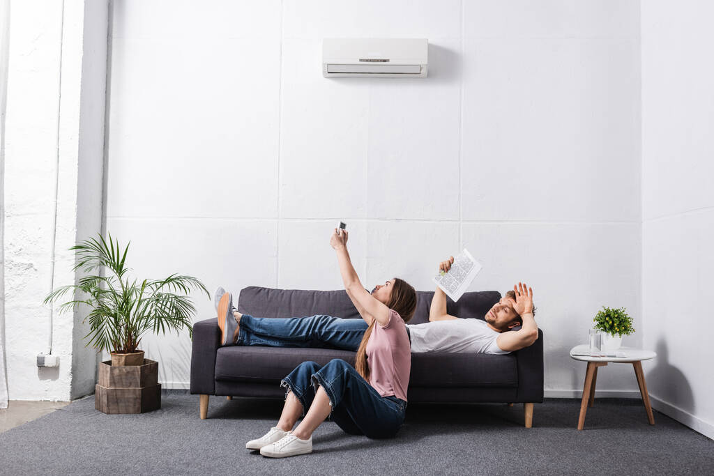 junges Paar mit Fernbedienung und Zeitung leidet unter Hitze, während es mit defekter Klimaanlage zu Hause sitzt  - Foto, Bild