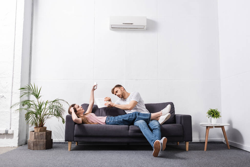 Trauriges junges Paar mit Fernbedienung leidet unter Hitze zu Hause mit defekter Klimaanlage  - Foto, Bild