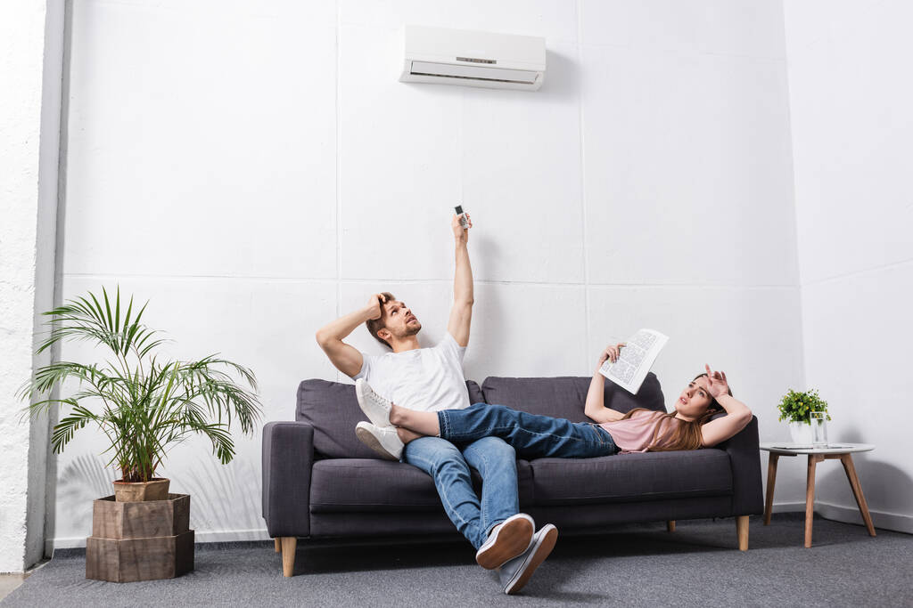 frustrated young couple with remote controller suffering from heat at home with broken air conditioner  - Photo, Image
