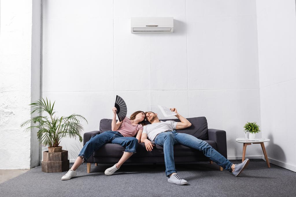 pareja joven con ventilador de mano y periódico que sufre de calor en casa con aire acondicionado roto
  - Foto, Imagen