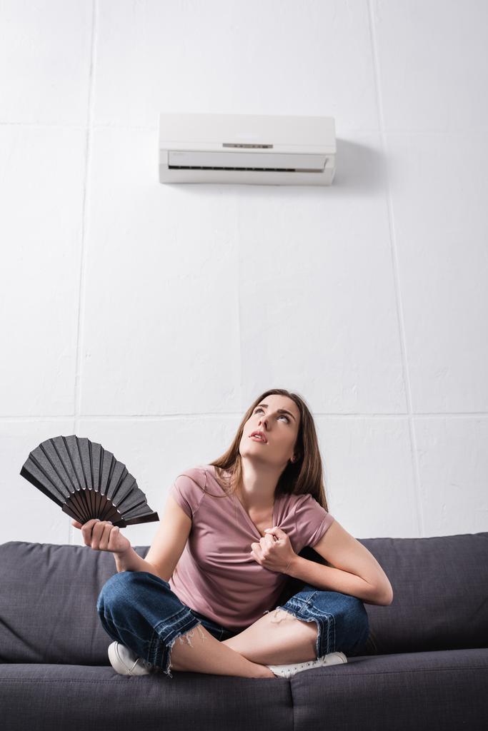 молодая женщина страдает от жары и с помощью ручного вентилятора дома со сломанным кондиционером
 - Фото, изображение