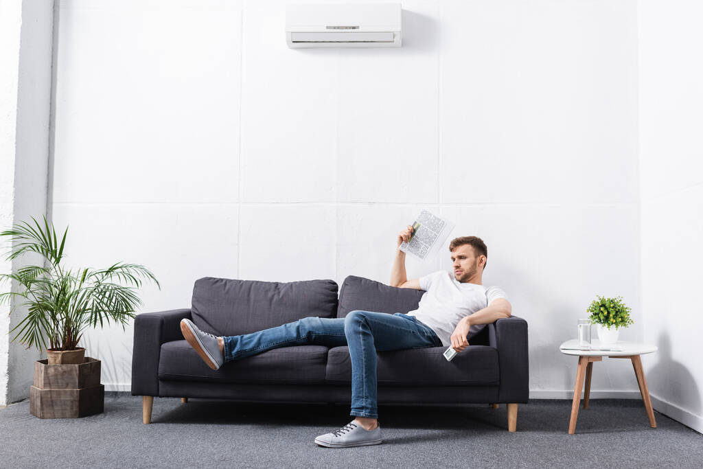 joven que sufre de calor mientras usa el periódico como ventilador de mano en casa con aire acondicionado roto
 - Foto, Imagen