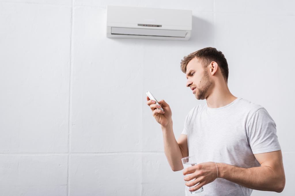 gefrustreerde man die glas water vasthoudt terwijl hij probeert de airconditioner aan te zetten met kapotte afstandsbediening thuis  - Foto, afbeelding