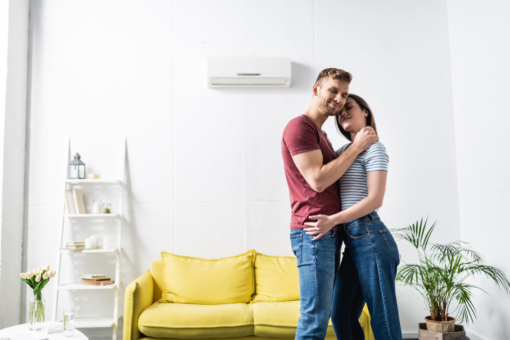 uśmiechnięta para przytulająca się w domu z klimatyzatorem - Zdjęcie, obraz