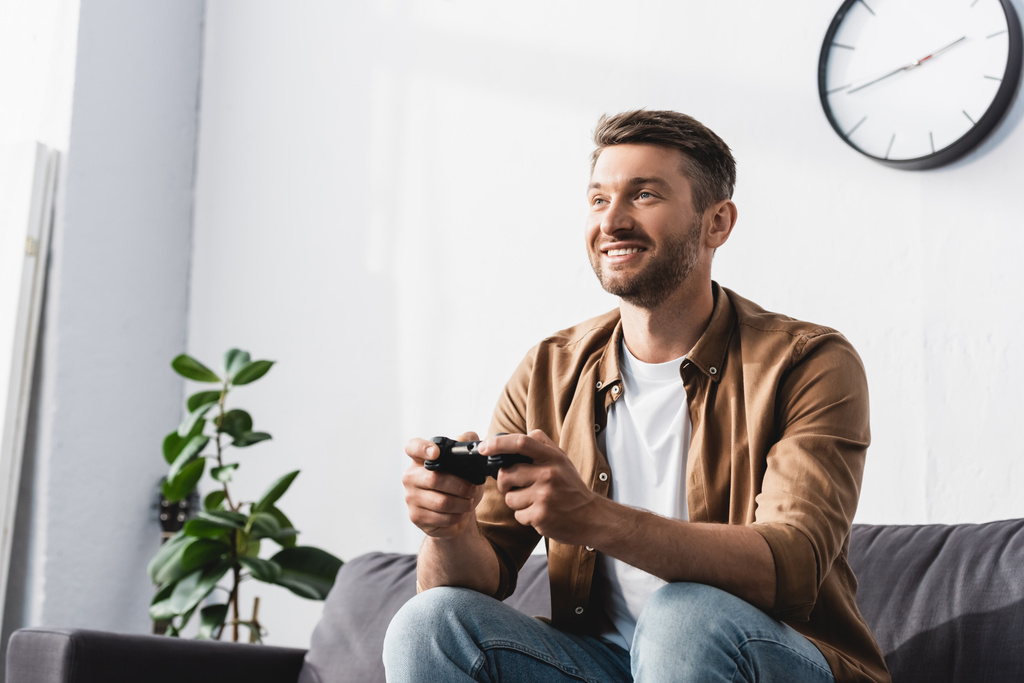 KYIV, UKRAINE - JUNE 9, 2020: cheerful man sitting on sofa and playing video game with joystick - Photo, Image