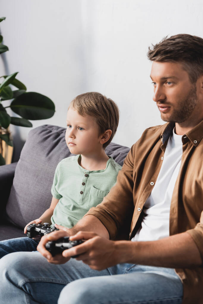 KYIV, UCRANIA - 9 de junio de 2020: padre e hijo concentrados sentados en el sofá y jugando videojuegos con joysticks
 - Foto, imagen