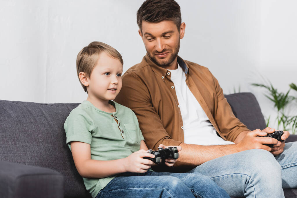 KYIV, UKRAINE - JUNE 9, 2020: happy father looking at smiling son while playing video game with joysticks - Photo, Image