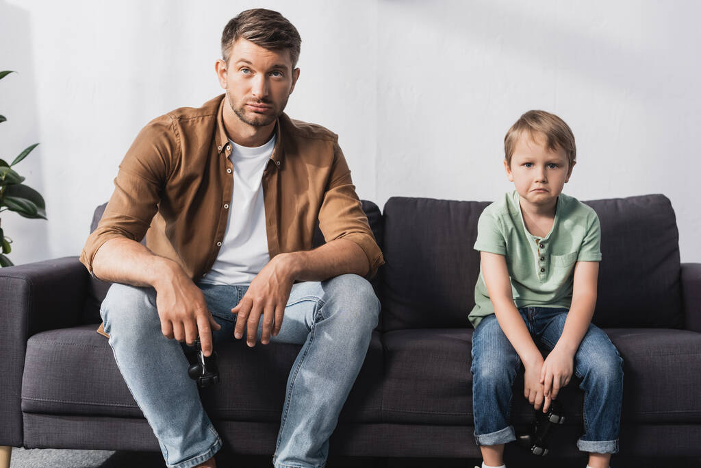 KYIV, UCRAINA - 9 GIUGNO 2020: padre e figlio sconvolti che guardano la macchina fotografica seduti sul divano con i joystick - Foto, immagini