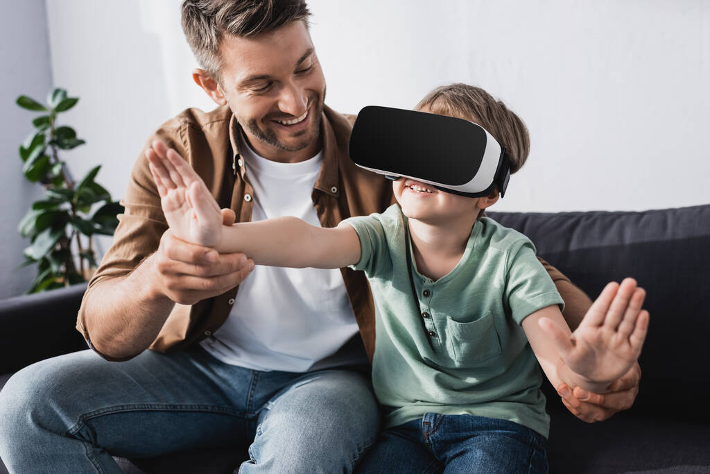 smiling man touching hands of cheerful son in vr headset  - Photo, Image