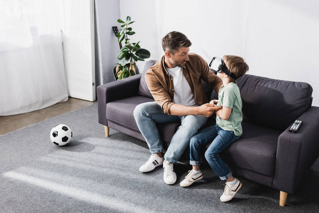высокий угол зрения мужчины, трогающего сына с помощью гарнитуры VR, сидя на диване
 - Фото, изображение