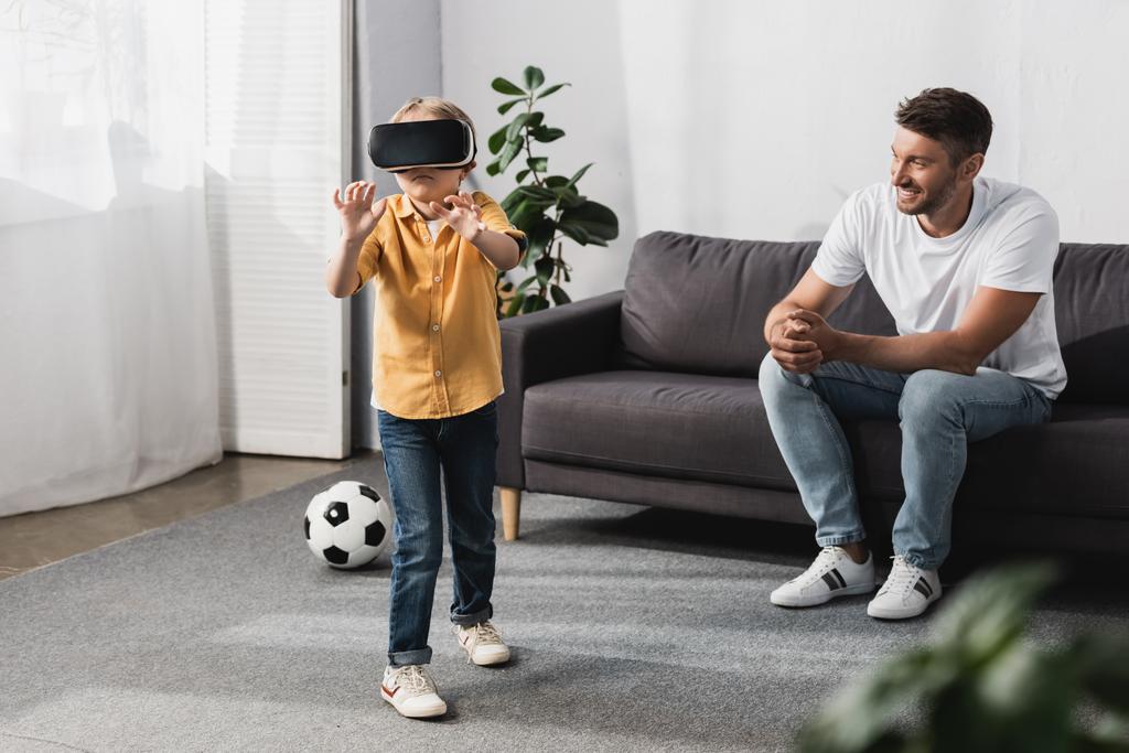 selective focus of cute boy standing and gesturing in vr headset near smiling father sitting on sofa - Photo, Image