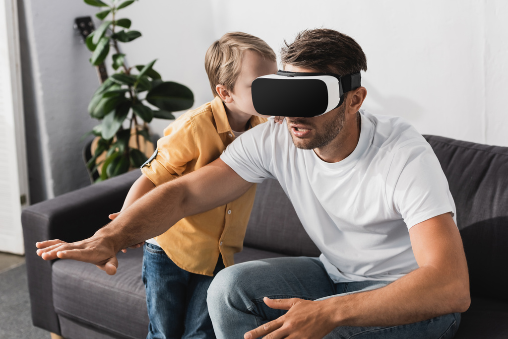 VR kulaklıklı heyecanlı bir adam uzanmış elleriyle oturuyor ve sevimli oğlu onunla konuşuyor. - Fotoğraf, Görsel