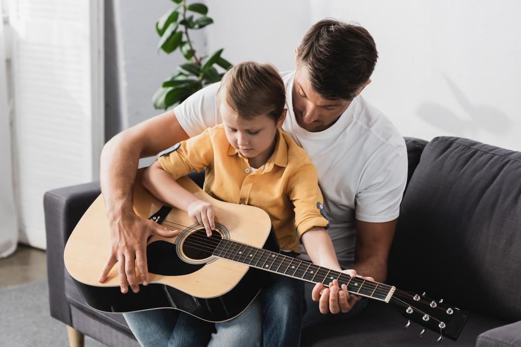 menino bonito sentado de joelhos pais e aprender a tocar guitarra acústica
 - Foto, Imagem