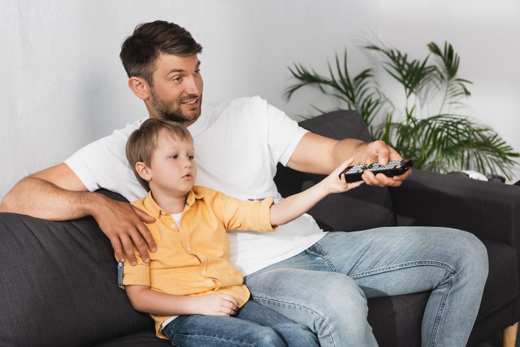 smiling father and bored son clicking channels with remote controller while watching tv - Photo, Image