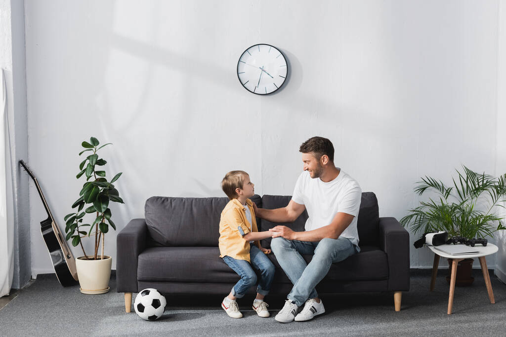 père et fils parlent assis sur le canapé près du ballon de football et des plantes en pot - Photo, image