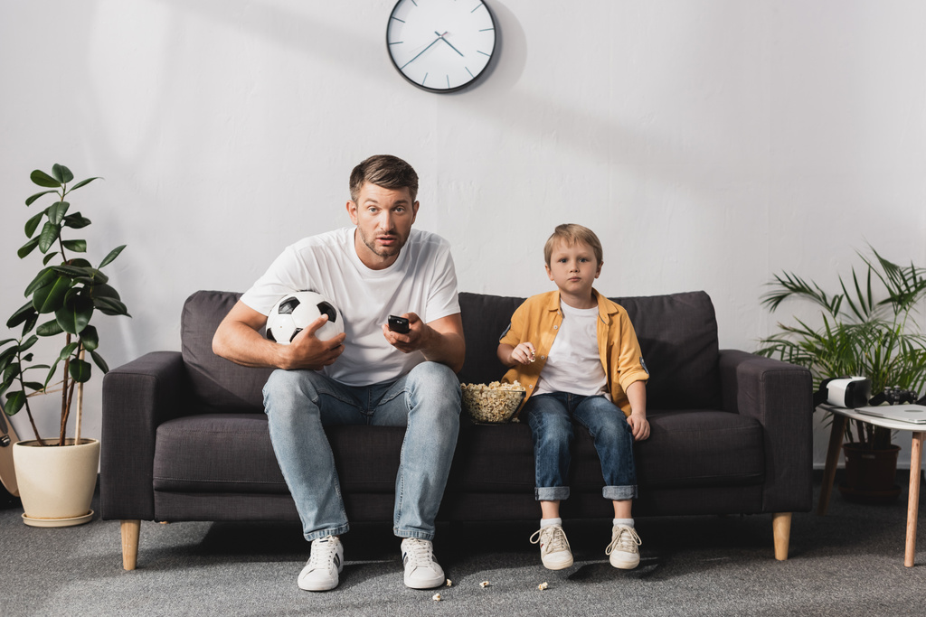 zmartwiony człowiek trzyma piłkę nożną i TV zdalne kontrolery w pobliżu syna jedząc popcorn - Zdjęcie, obraz