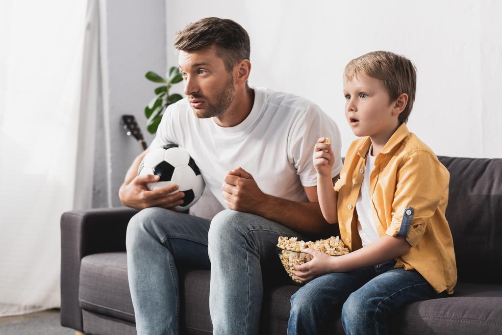 стурбований батько і син дивиться телевізор, тримаючи футбольний м'яч і попкорн
 - Фото, зображення