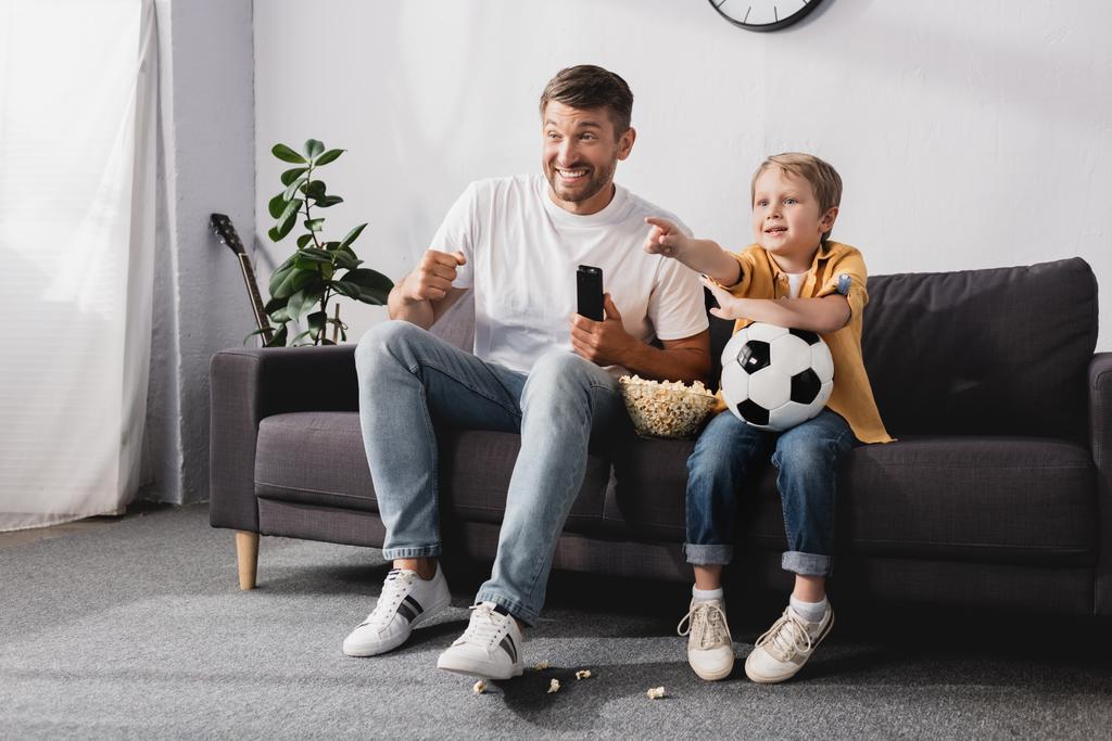 niño feliz sosteniendo la pelota de fútbol y señalando con el dedo cerca padre emocionado mientras mira la televisión
 - Foto, imagen