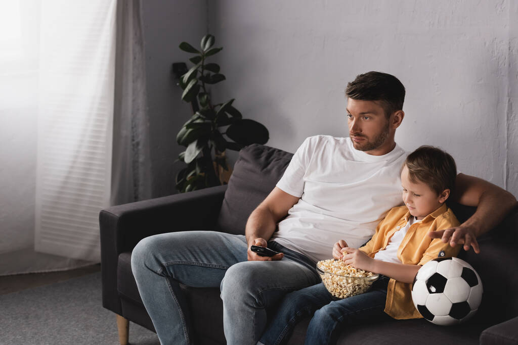 退屈した父と息子サッカーボールの近くにポップコーンのボウルとテレビを見て - 写真・画像