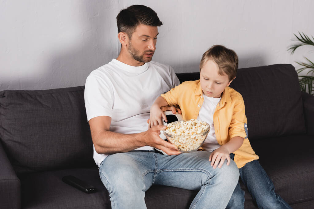 ojciec i syn jedzą popcorn siedząc na kanapie i oglądając telewizję - Zdjęcie, obraz