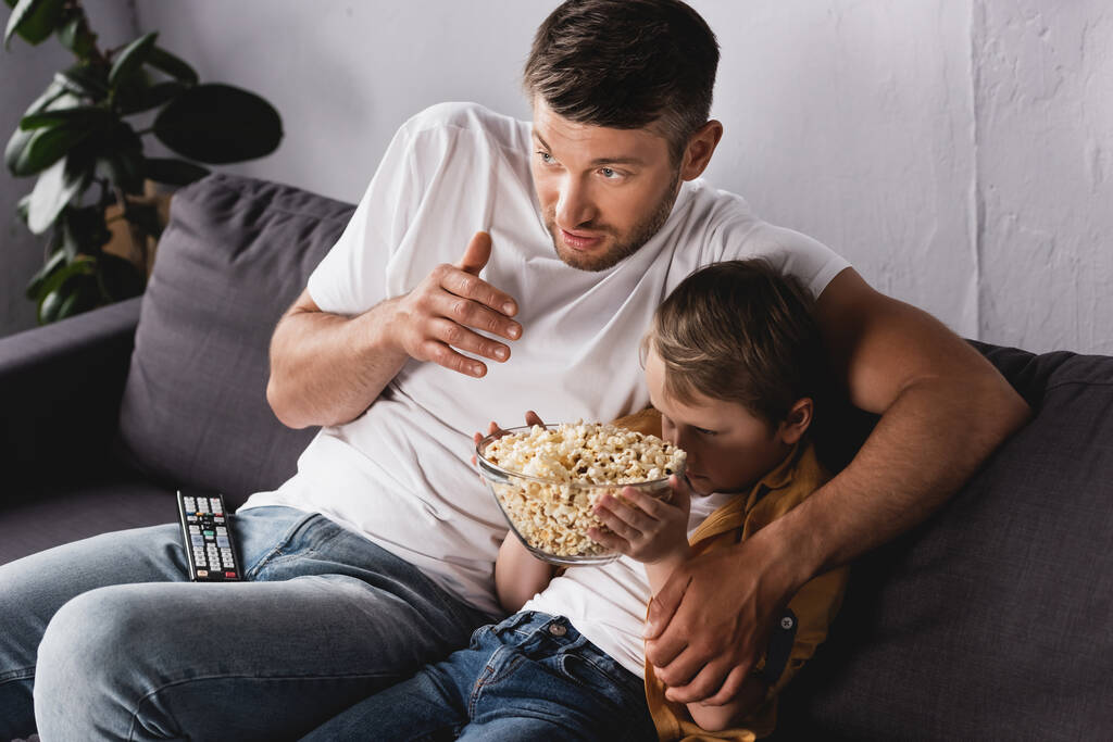 Konzentrierter Vater neben verängstigtem Sohn, der sich vor dem Fernseher mit einer Schüssel Popcorn versteckt - Foto, Bild