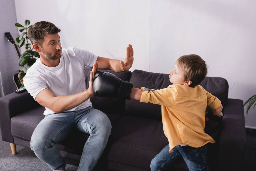 χαριτωμένο αγόρι σε γάντια πυγμαχίας αγωνίζονται με τον πατέρα κάθεται στον καναπέ - Φωτογραφία, εικόνα