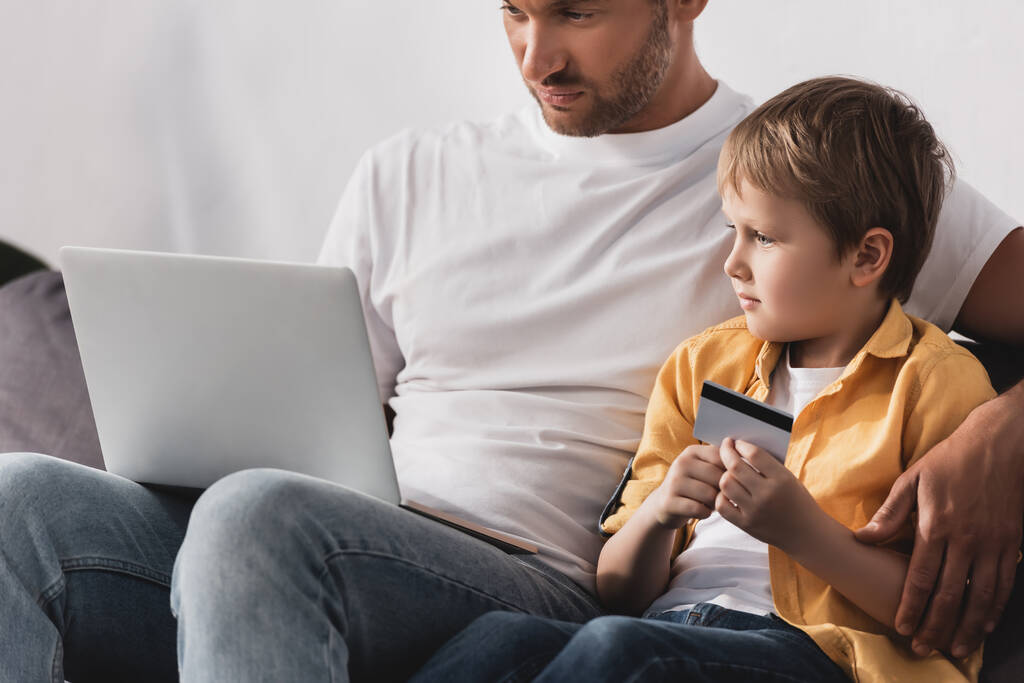 συμπυκνωμένο πατέρα χρησιμοποιώντας φορητό υπολογιστή, ενώ αξιολάτρευτο γιο κρατώντας πιστωτική κάρτα - Φωτογραφία, εικόνα