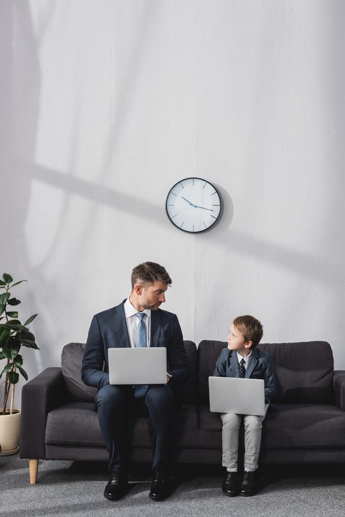 vakava liikemies ja hänen poikansa virallisessa kulumista istuu sohvalla kannettavat tietokoneet ja katselevat toisiaan - Valokuva, kuva
