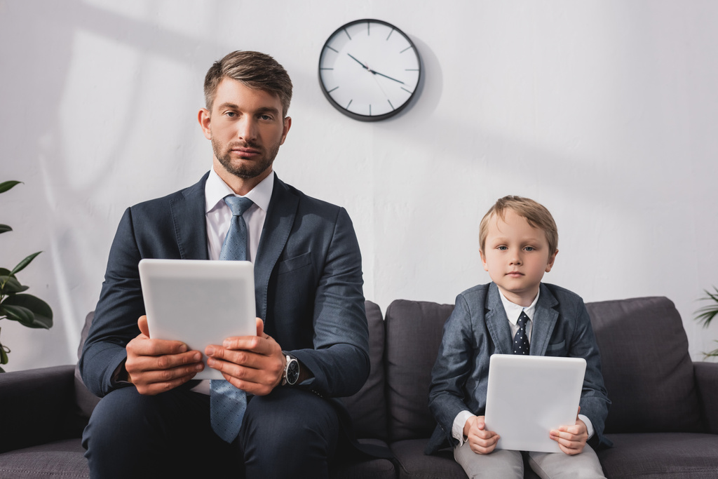 seriózní podnikatel a jeho syn ve formálním oblečení drží digitální tablety, zatímco sedí na pohovce doma - Fotografie, Obrázek