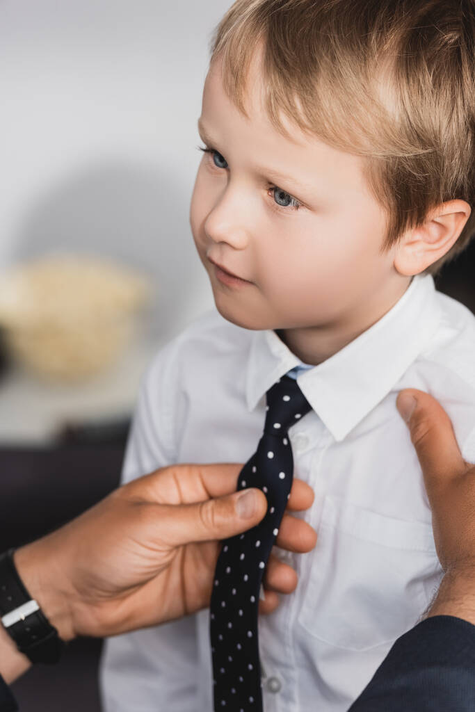 cropped view of businessman putting tie on adorable, serious son at home - Photo, Image