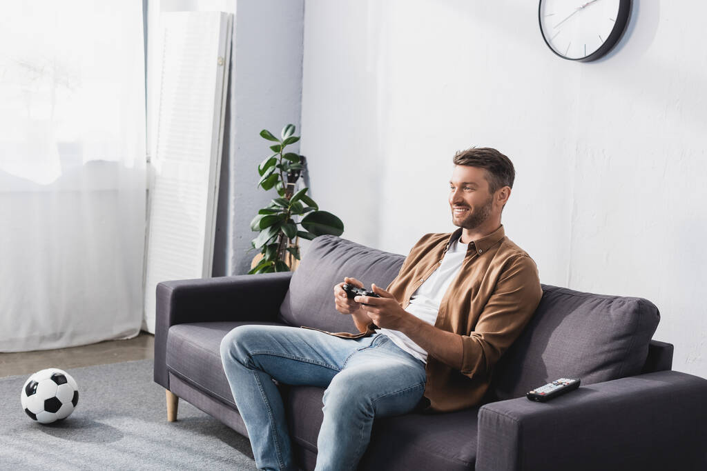 KYIV, UCRANIA - 9 de junio de 2020: hombre alegre jugando videojuegos con joystick mientras está sentado en el sofá cerca de la pelota de fútbol
 - Foto, imagen