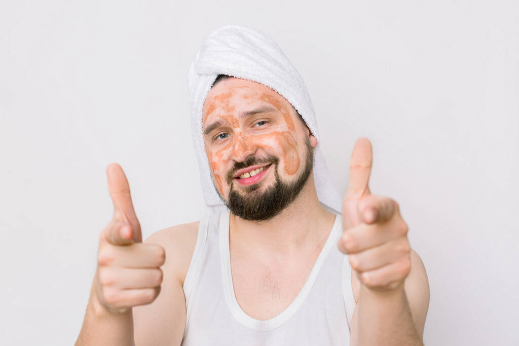 Πορτρέτο ενός όμορφου χαμογελαστού νεαρού με μάσκα από λάσπη προσώπου, που δείχνει τα δάχτυλά του στην κάμερα. Περιποίηση προσώπου και σώματος για άνδρες. Απομονωμένα σε λευκό - Φωτογραφία, εικόνα