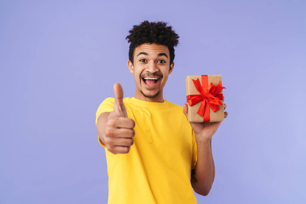 Фото счастливого африканского американца, держащего подарочную коробку и показывающего большой палец на фиолетовом фоне
 - Фото, изображение