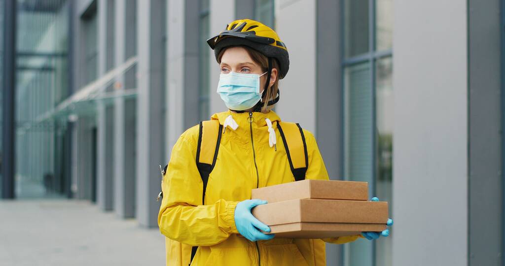 Portrét krásné ženy doručovatelské rukavice v lékařské masce stojící na ulici a držící kartónové krabice. Krásná kurýrka s balíčky - Fotografie, Obrázek