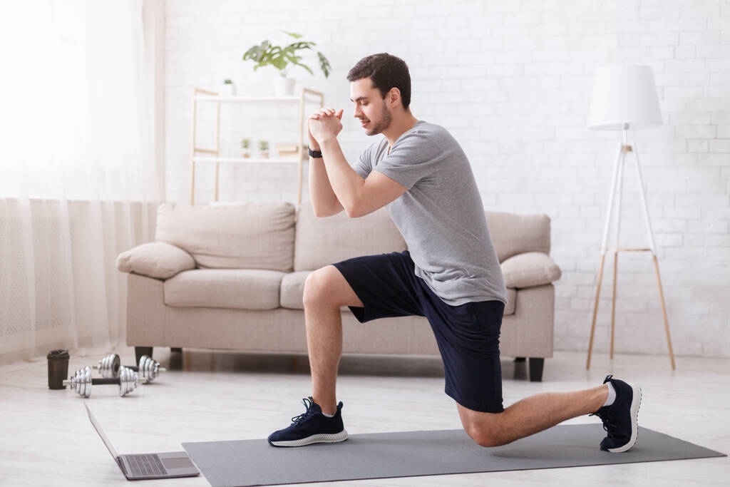Αθλητικές δραστηριότητες στο σπίτι. Νεαρός άνδρας παρακολουθεί online ασκήσεις και κάνει lunges - Φωτογραφία, εικόνα
