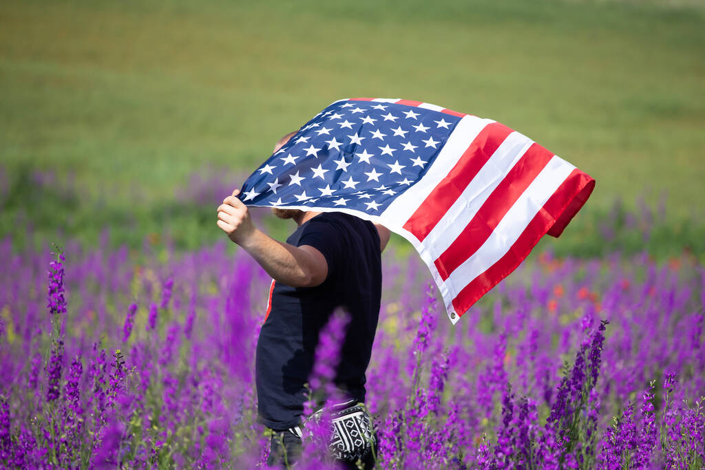 Ελκυστικός άνδρας με σημαία των Ηνωμένων Πολιτειών σε ένα όμορφο καλοκαιρινό πεδίο σε μια σαφή, ηλιόλουστη μέρα. Γιορτάζοντας την Ημέρα της Ανεξαρτησίας, Εθνική ιδέα γιορτή. - Φωτογραφία, εικόνα