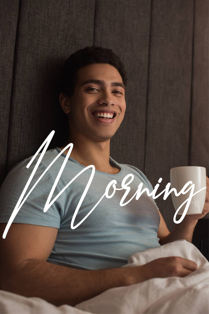 glimlachende gemengde ras man met kopje koffie in de buurt ochtend belettering in de slaapkamer  - Foto, afbeelding