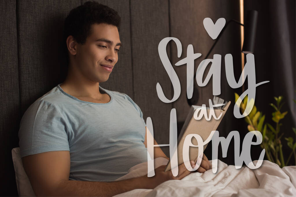 smiling mixed race man reading book near stay at home lettering in bedroom  - Photo, Image