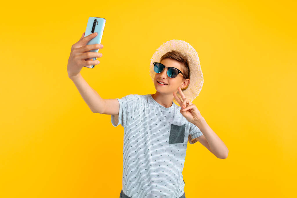 Boldog mosolygós tini srác egy nyári kalap csinál egy szelfi egy mobiltelefon, egy elszigetelt sárga háttér - Fotó, kép