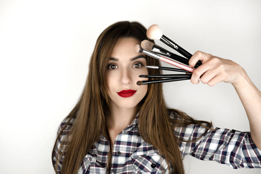 brunette young woman with red lips holding makeup brushes in one hand near face , beauty and style, isolated white background. - Photo, Image