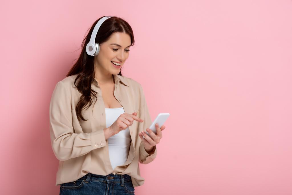 Positive Frau mit Kopfhörer zeigt mit dem Finger auf Smartphone auf rosa Hintergrund - Foto, Bild
