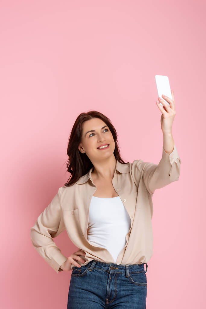 Attraktive Frau lächelt beim Selfie mit Smartphone auf rosa Hintergrund - Foto, Bild