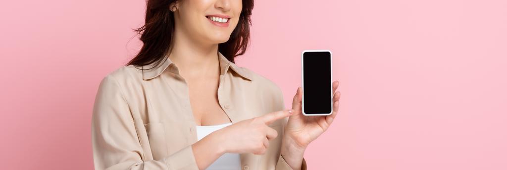 Panoramatická úroda usmívající se ženy ukazující prstem na smartphone izolovaný na růžové  - Fotografie, Obrázek